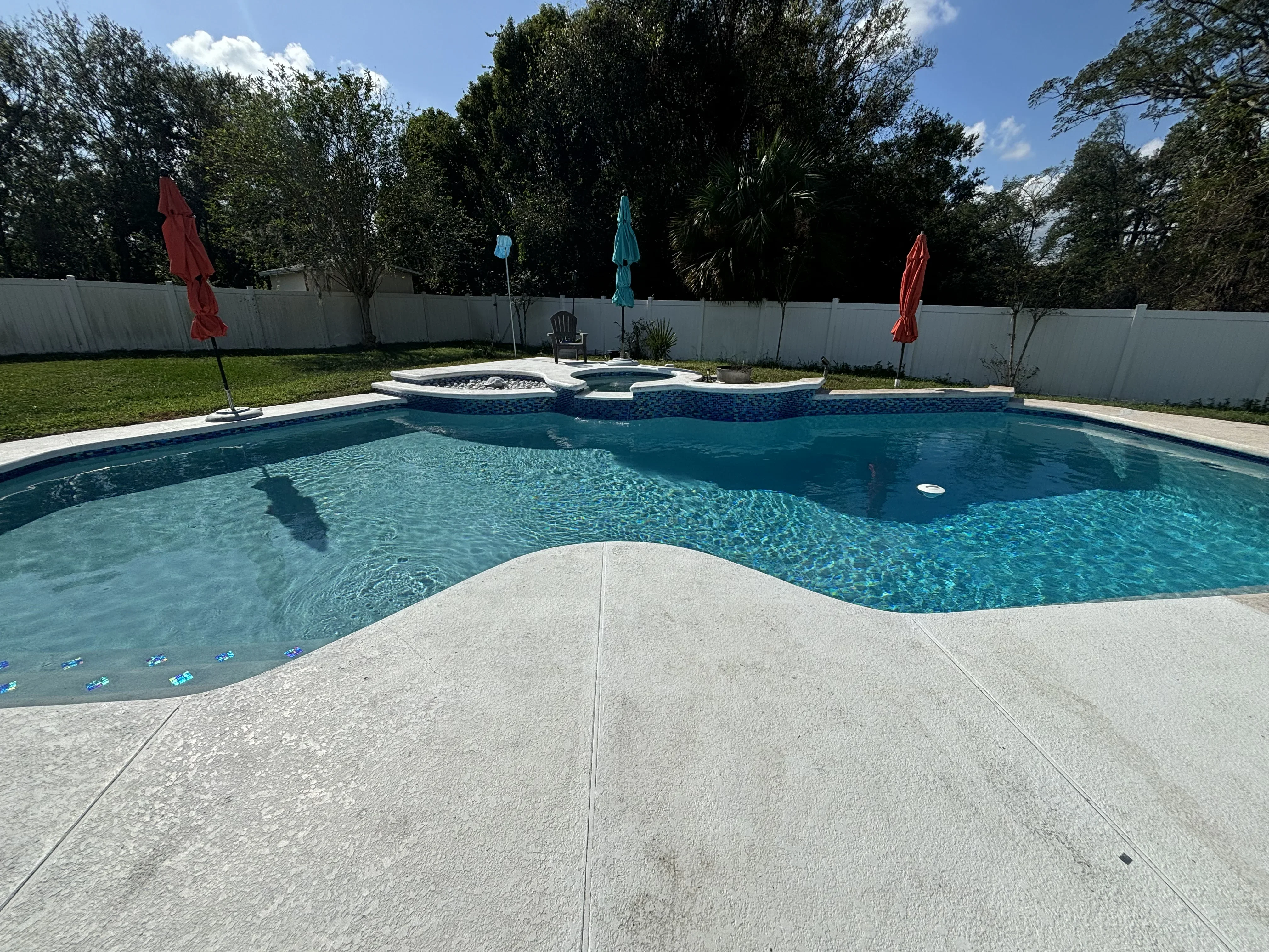 Freeform pool with spa, concrete deck, and white vinyl fence in landscaped backyard