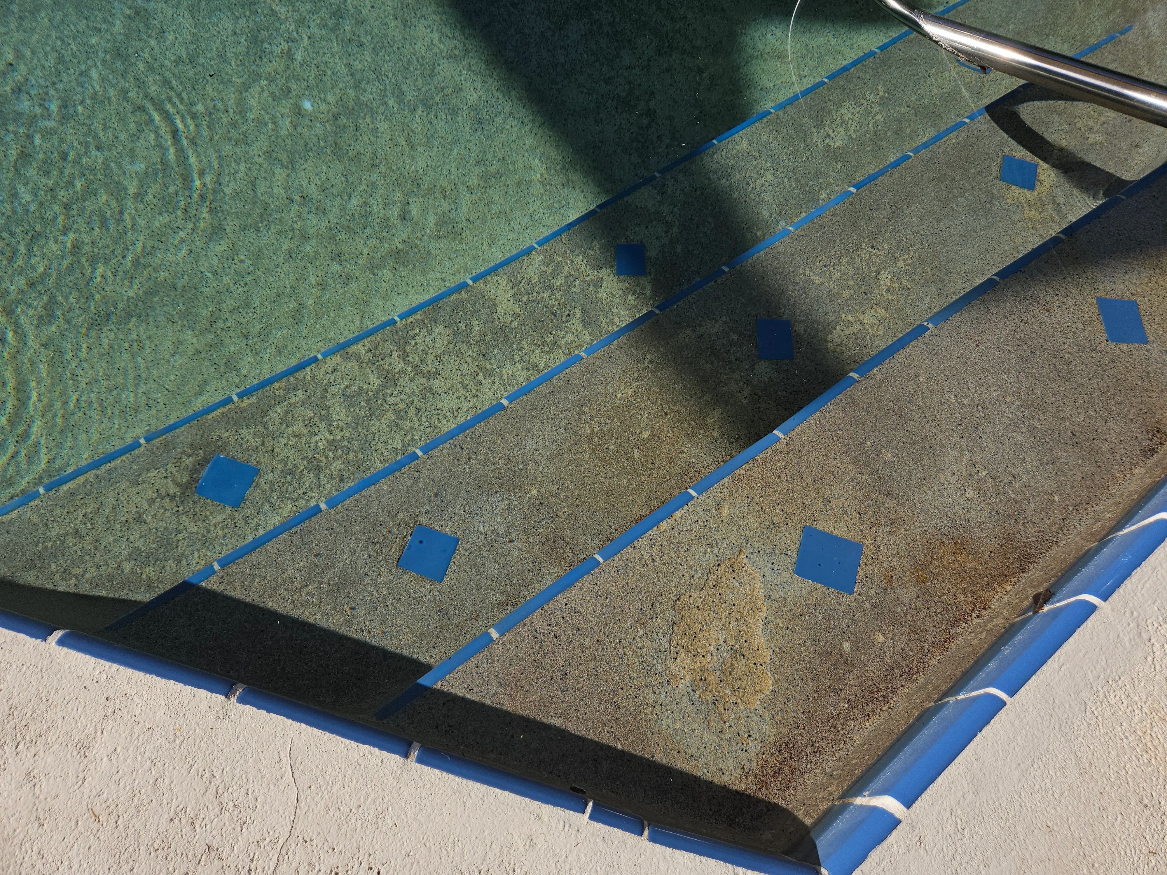 Pool floor cleaning in progress with blue tape sections and algae removal on concrete surface