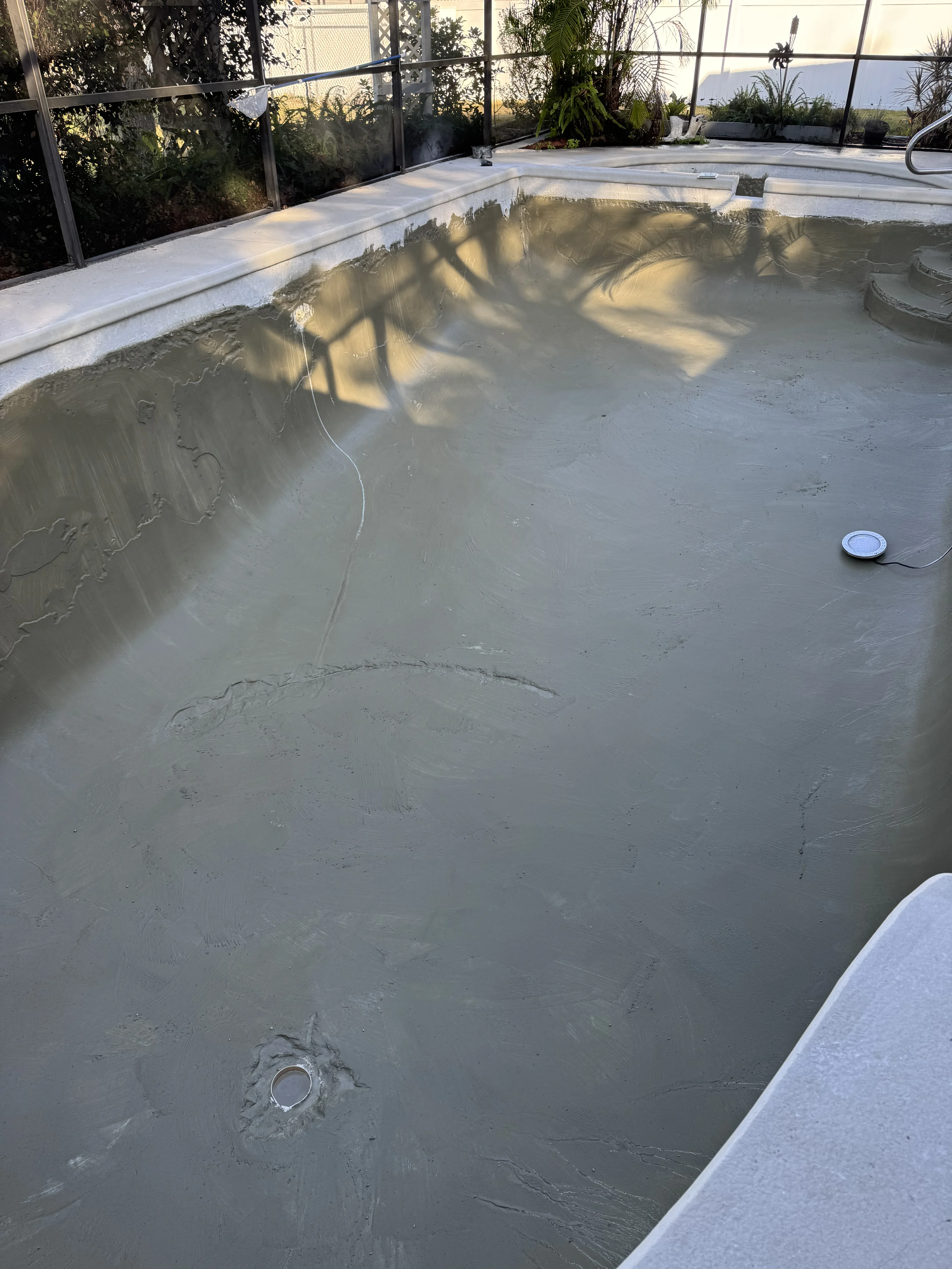 Drained pool basin with concrete floor, drain fixtures, and white deck surround in screened enclosure