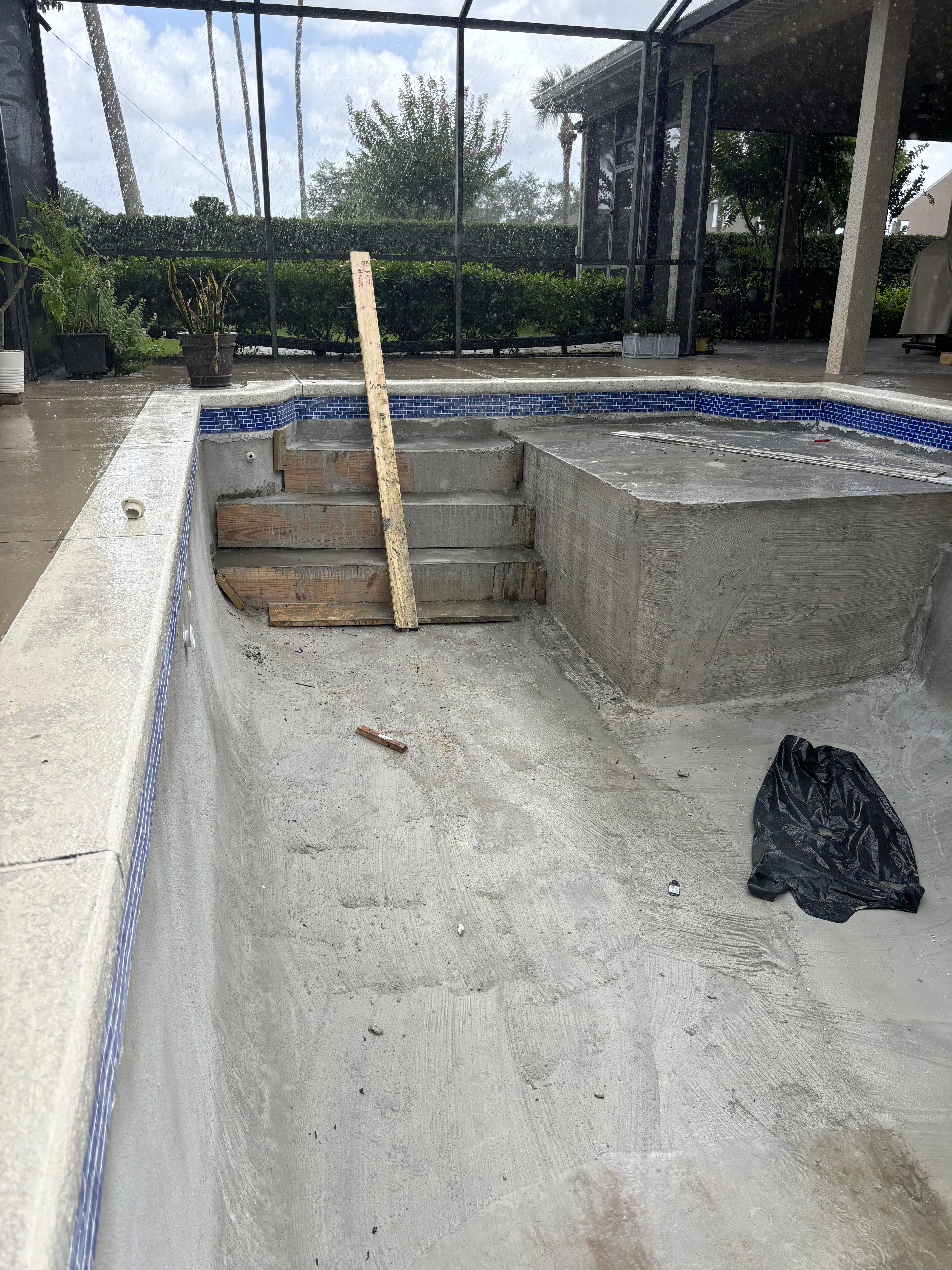 Drained pool basin showing concrete floor and walls with blue tile trim, wooden support structures, and screened enclosure