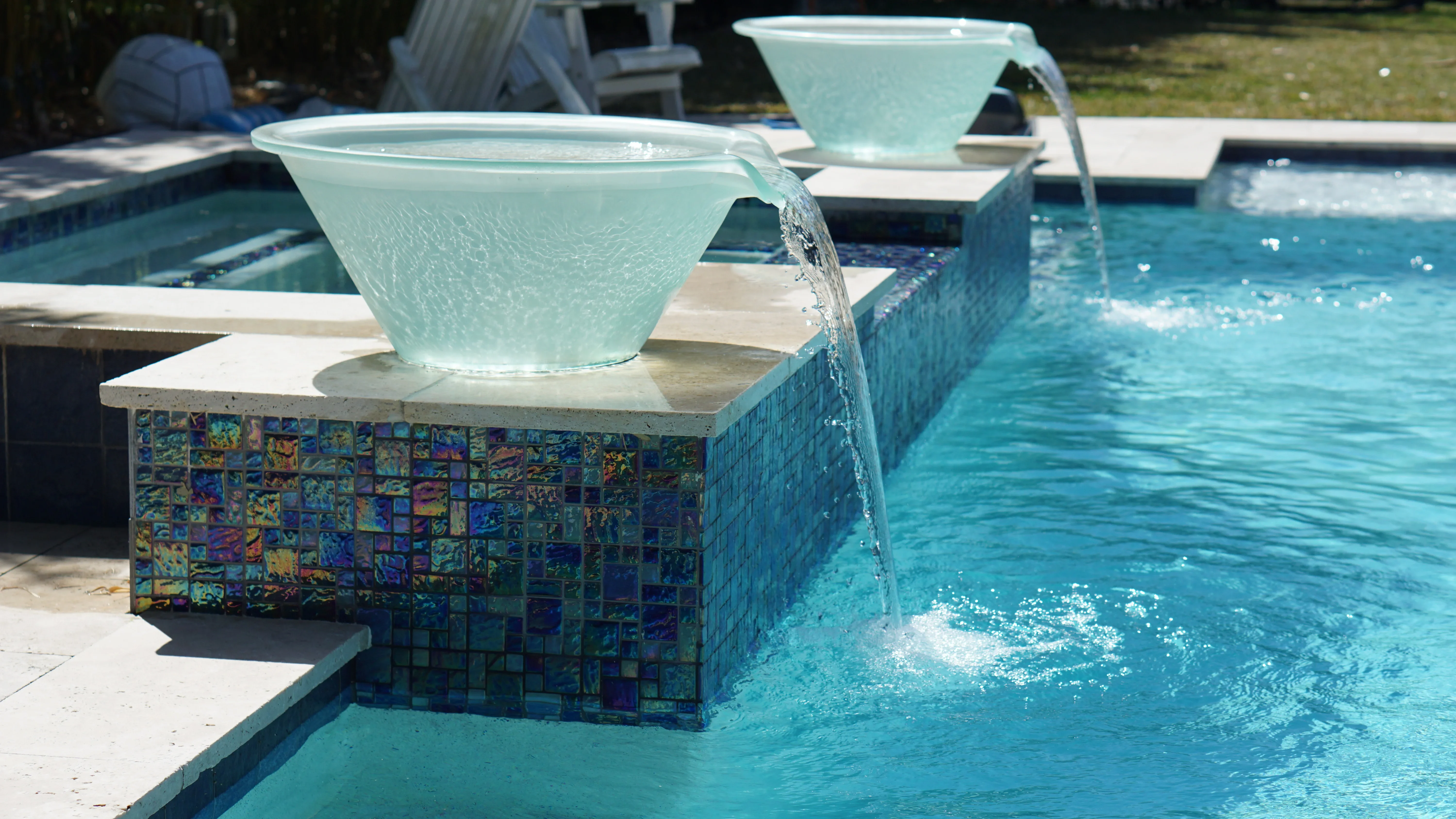Glass bowl water feature with cascading waterfall and decorative mosaic tile base beside swimming pool