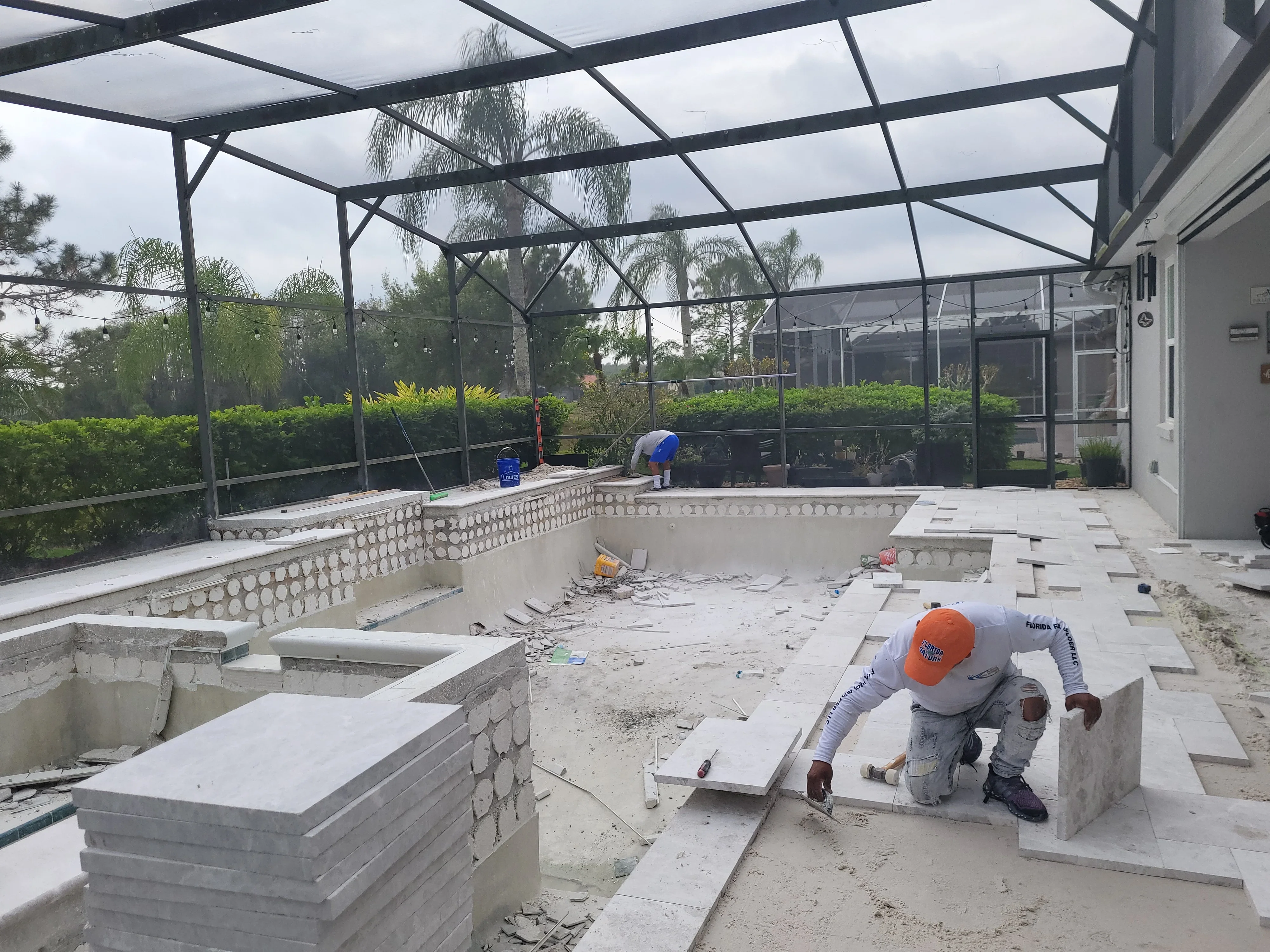 Worker installing concrete pool deck coping and pavers around in-ground pool with screen enclosure