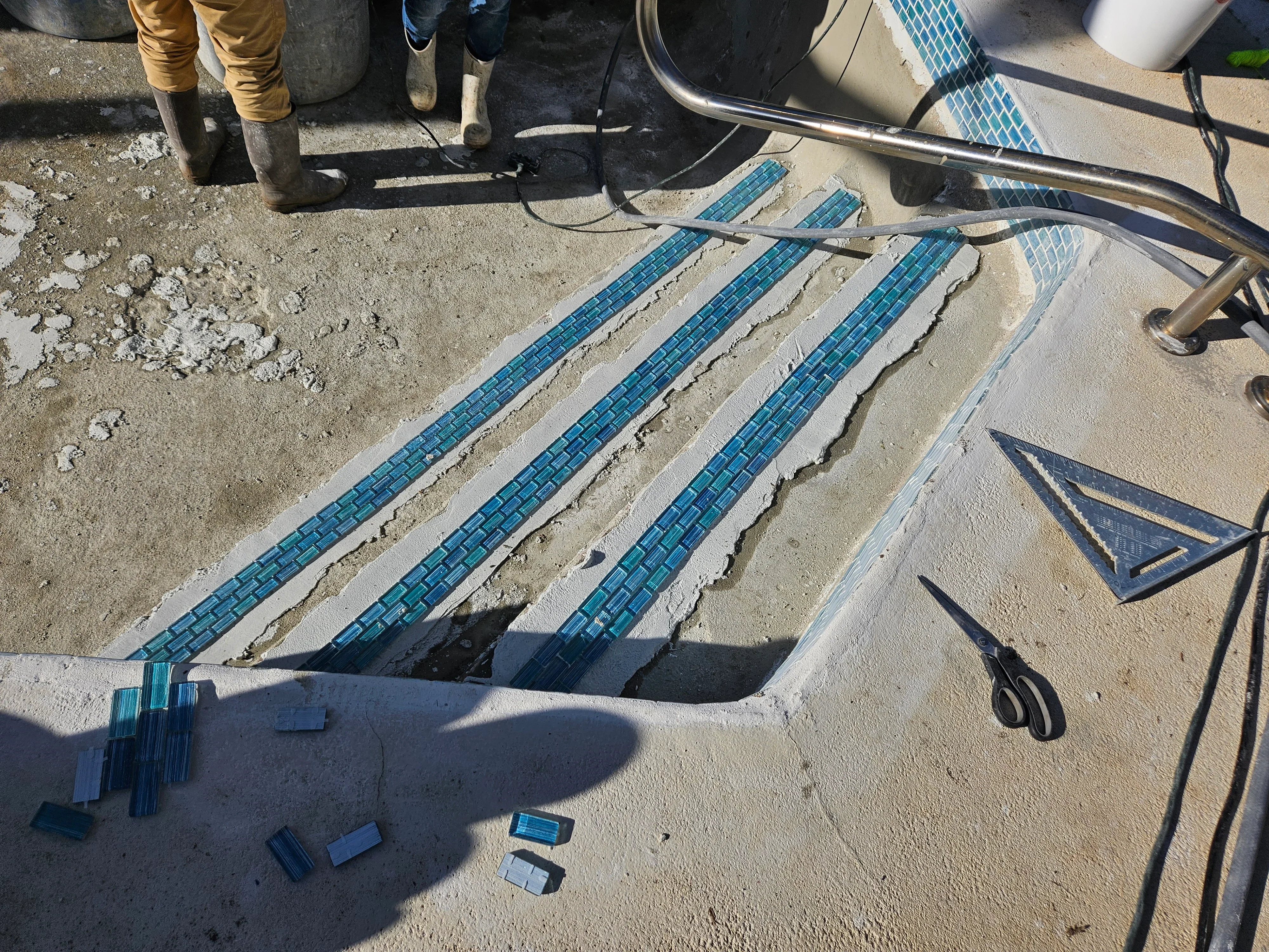 Blue ceramic pool deck tiles installed in three parallel rows on concrete with grout lines and installation tools
