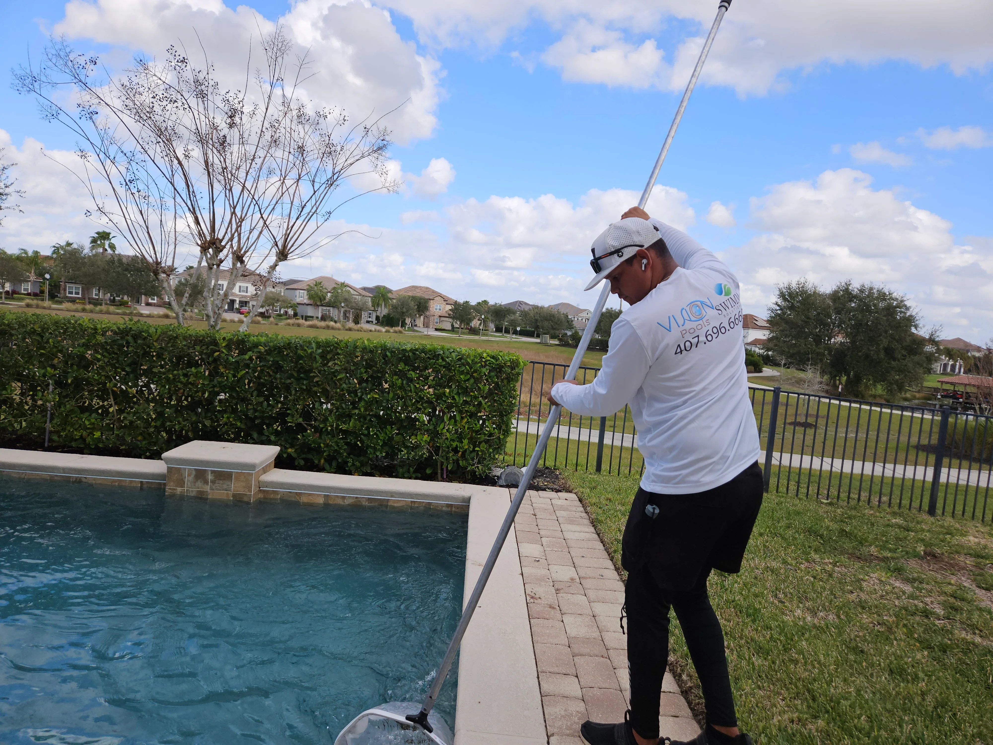 Technician performing pool maintenance with skimmer net on residential in-ground pool with brick deck
